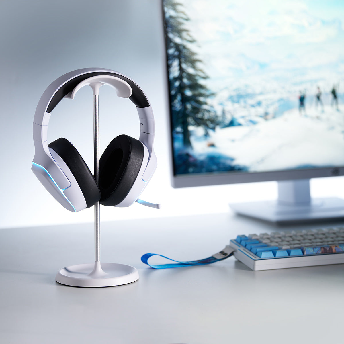 White gaming headset with blue LED accents on a stand. Monitor displays winter scene, keyboard visible.