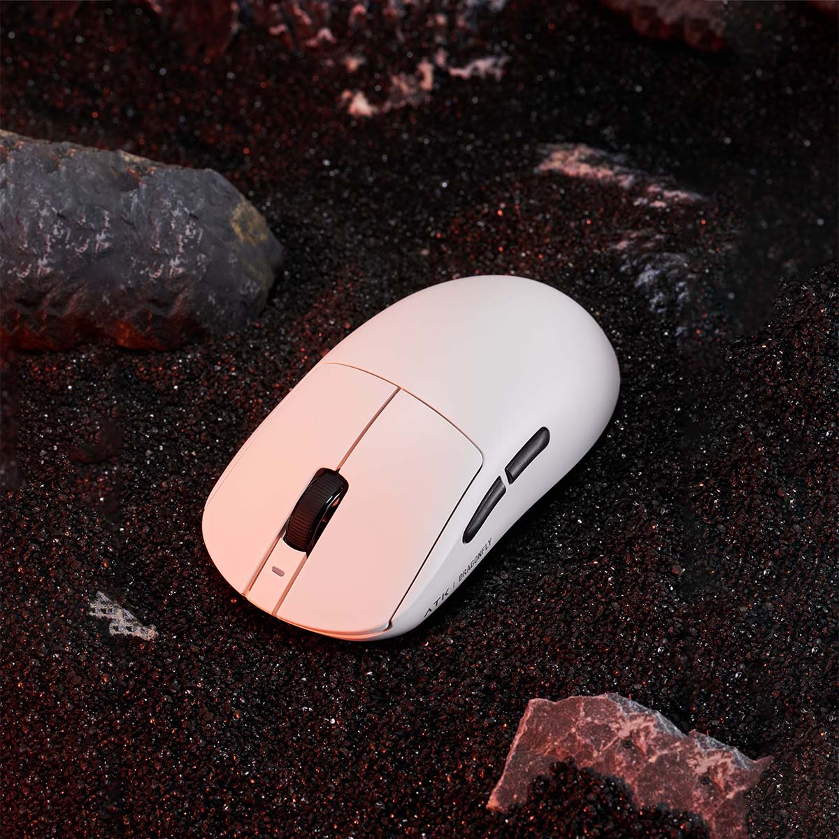 White ATK Dragonfly wireless gaming mouse on black sand with rocks.