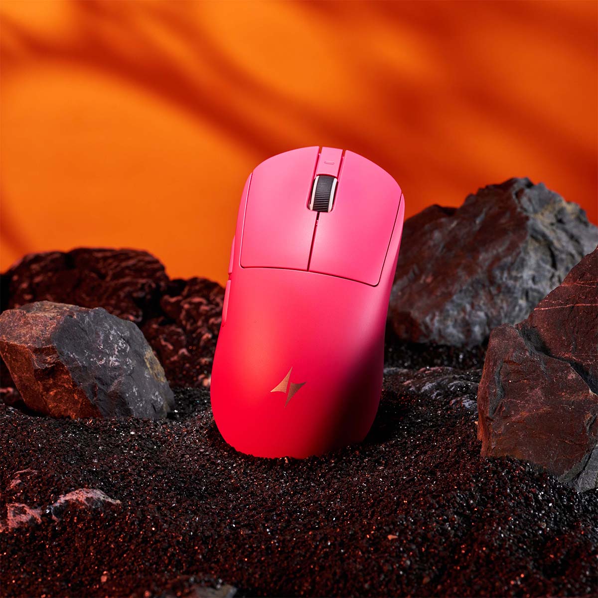 Pink ATK Dragonfly A9 gaming mouse on black sand with rocks. Dramatic orange background.